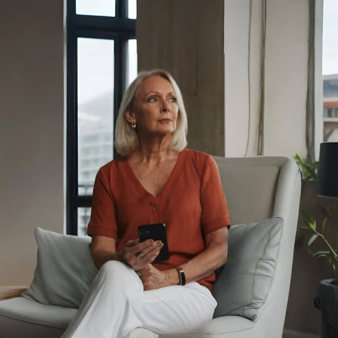 Older woman with shoulder-length gray hair sitting in a modern armchair, holding a smartphone and looking thoughtfully out the window