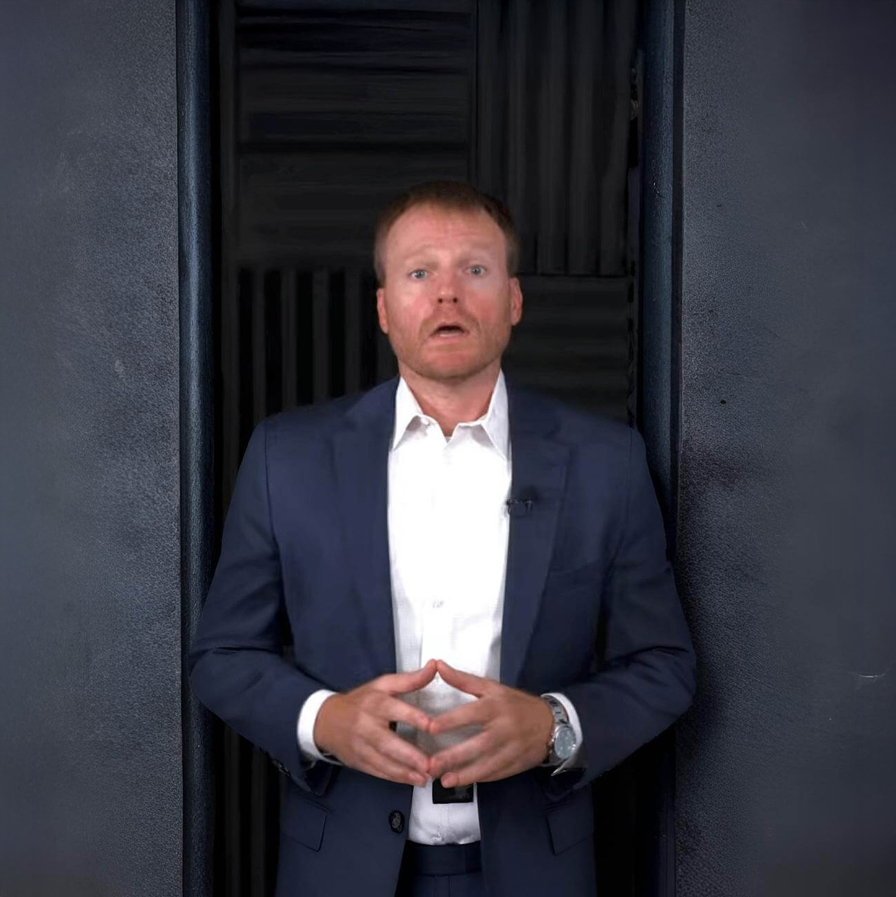 Chris Kelly in a navy suit and white shirt standing against a dark acoustic panel background